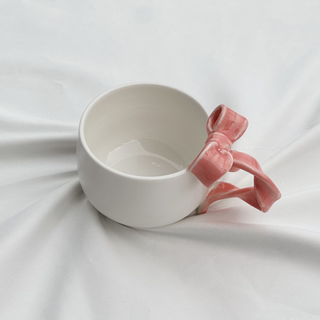 White mug with a pale pink twisted bow handle, sat on top of a white sheet.