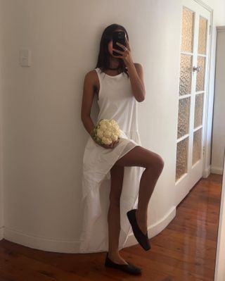 Woman wearing white halter dress with poplin skirt, black square-toed flats, holding cauliflower leaning against white wall