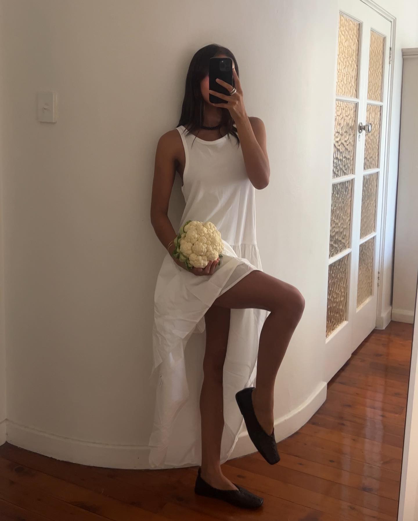 woman wearing white mixed media tank dress with poplin skirt, black square-toe flats, holding a cauliflower against white wall