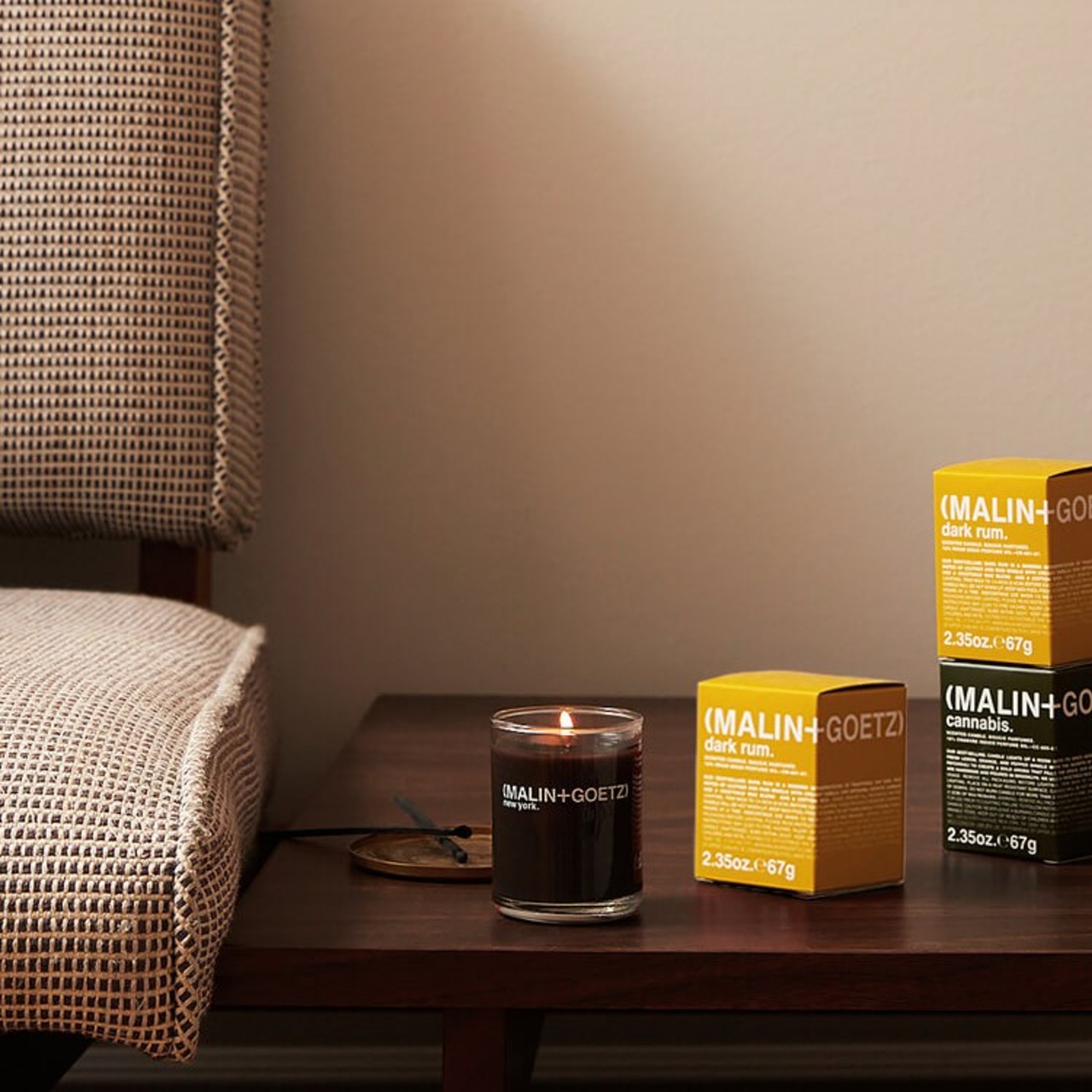 A lit Dark Rum scented candle from Malin + Goetz on a side table next to a sofa