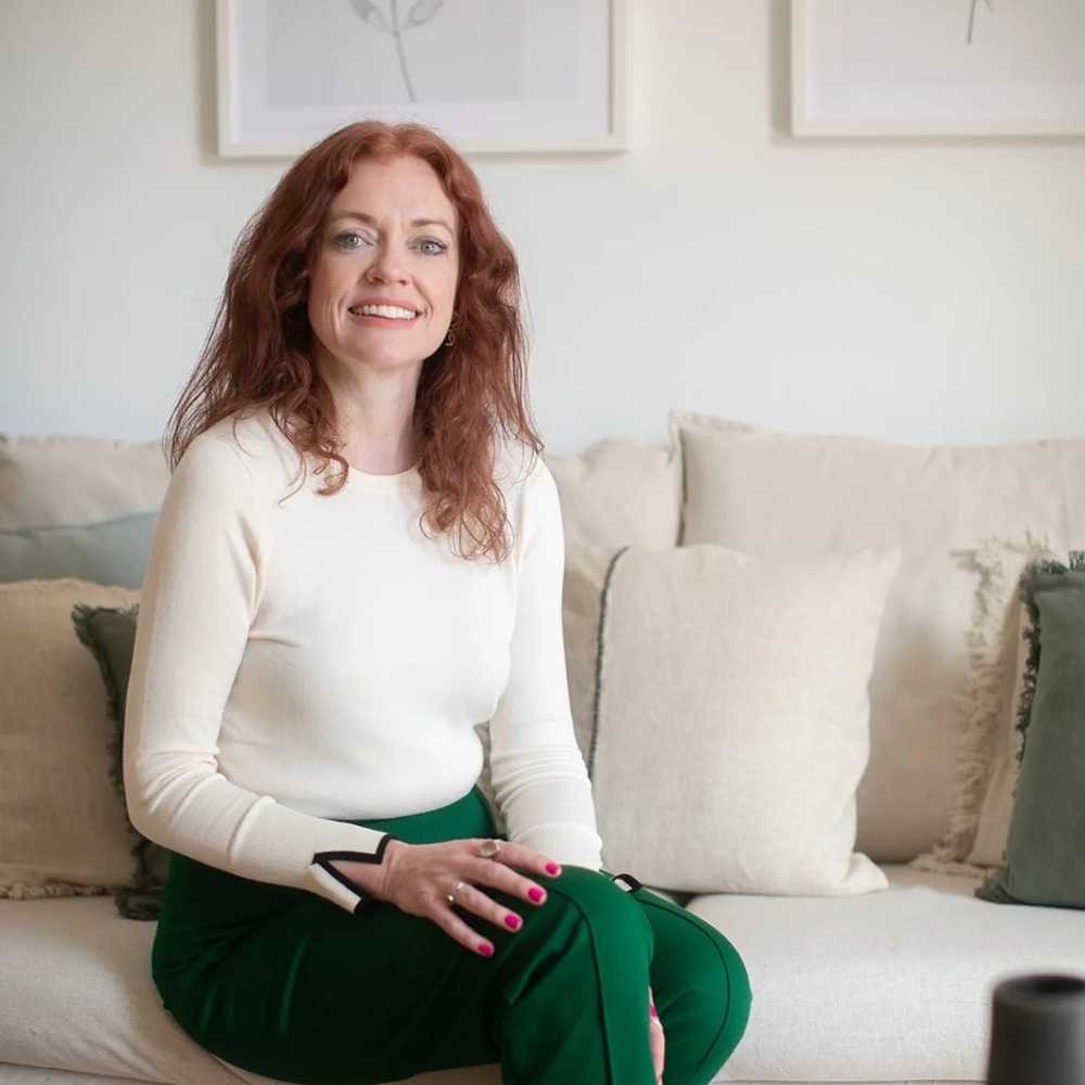female with long red hair, smiling and wearing cream top and green trousers sat on cream sofa