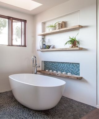 A corner of a white bathroom with three light wooden shelves with plants, candles, and jars on, a freestanding white curved bathtub with a silver tap, a floor with gray stones, and a window with wooden brown borders