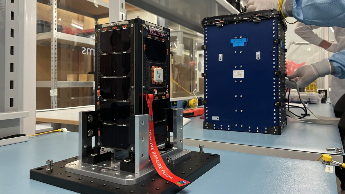 two cubesats sit on a table, gloved hands can be seen working on the farther.