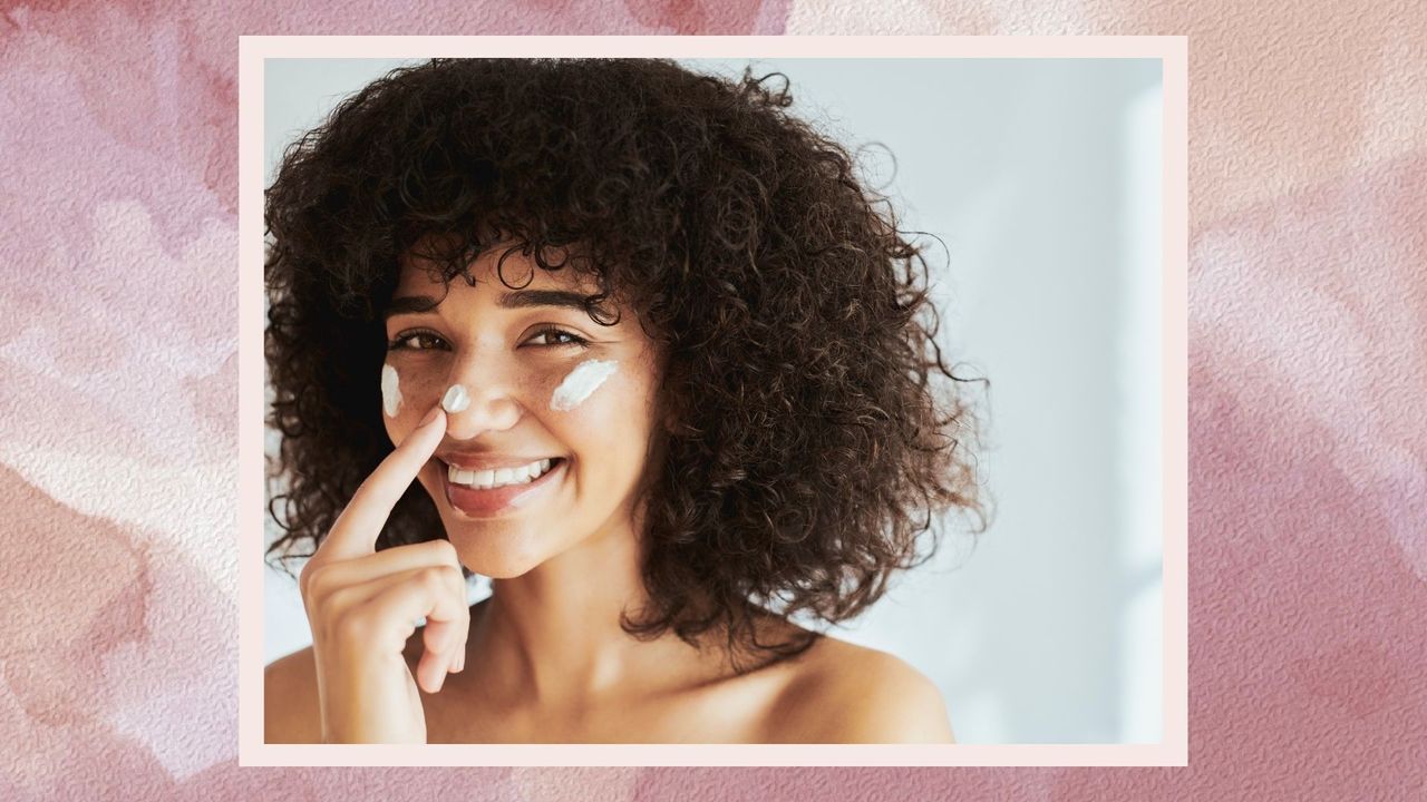 A woman applying sunscreen to her cheeks and nose while smiling on a pink watercolour backdrop