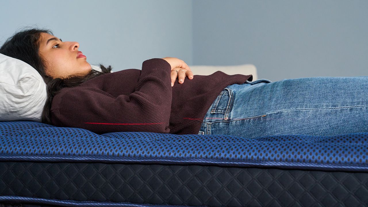 The image shows a dark haired mattress tester lying on her back on a dark blue mattress. Along her side, a laser level marks how deeply her spine sinks into the bed.