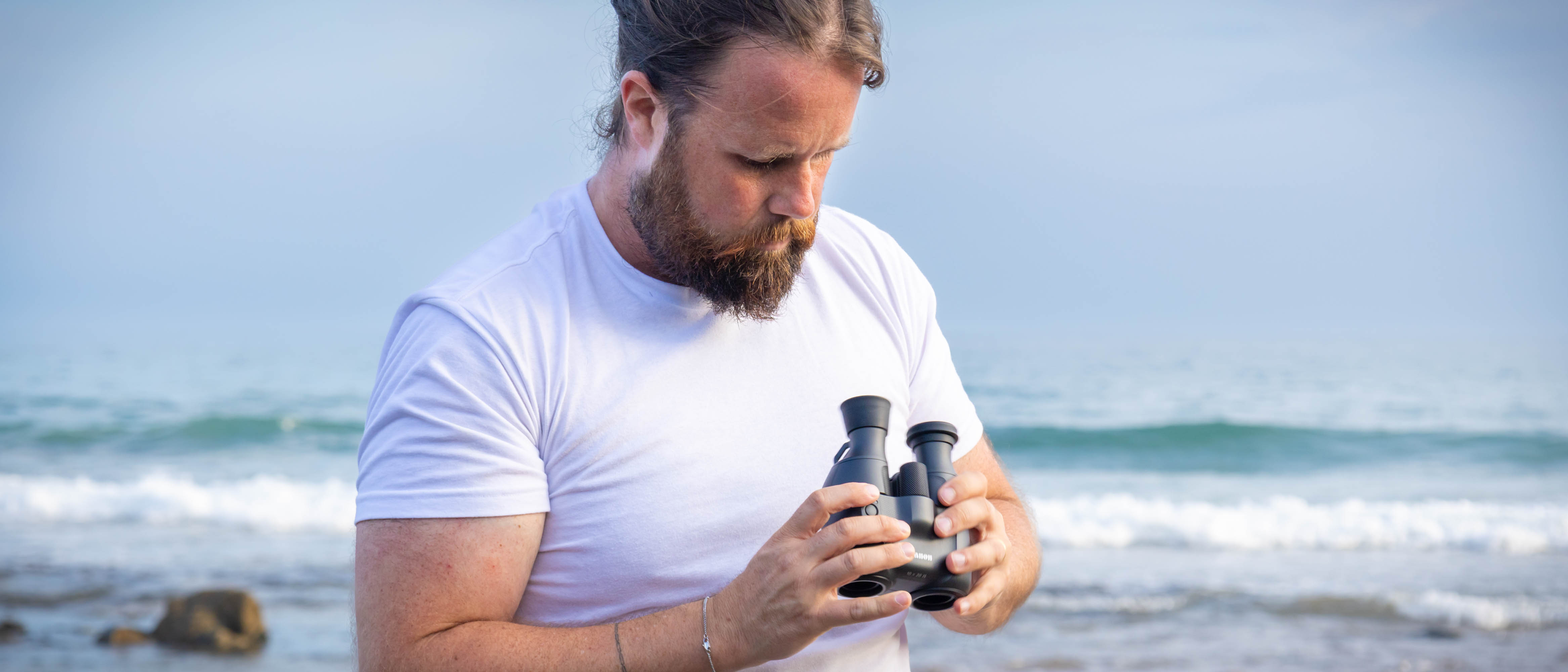A male looking at the Canon 10x20 IS binoculars on a beach, with the sea behind them.