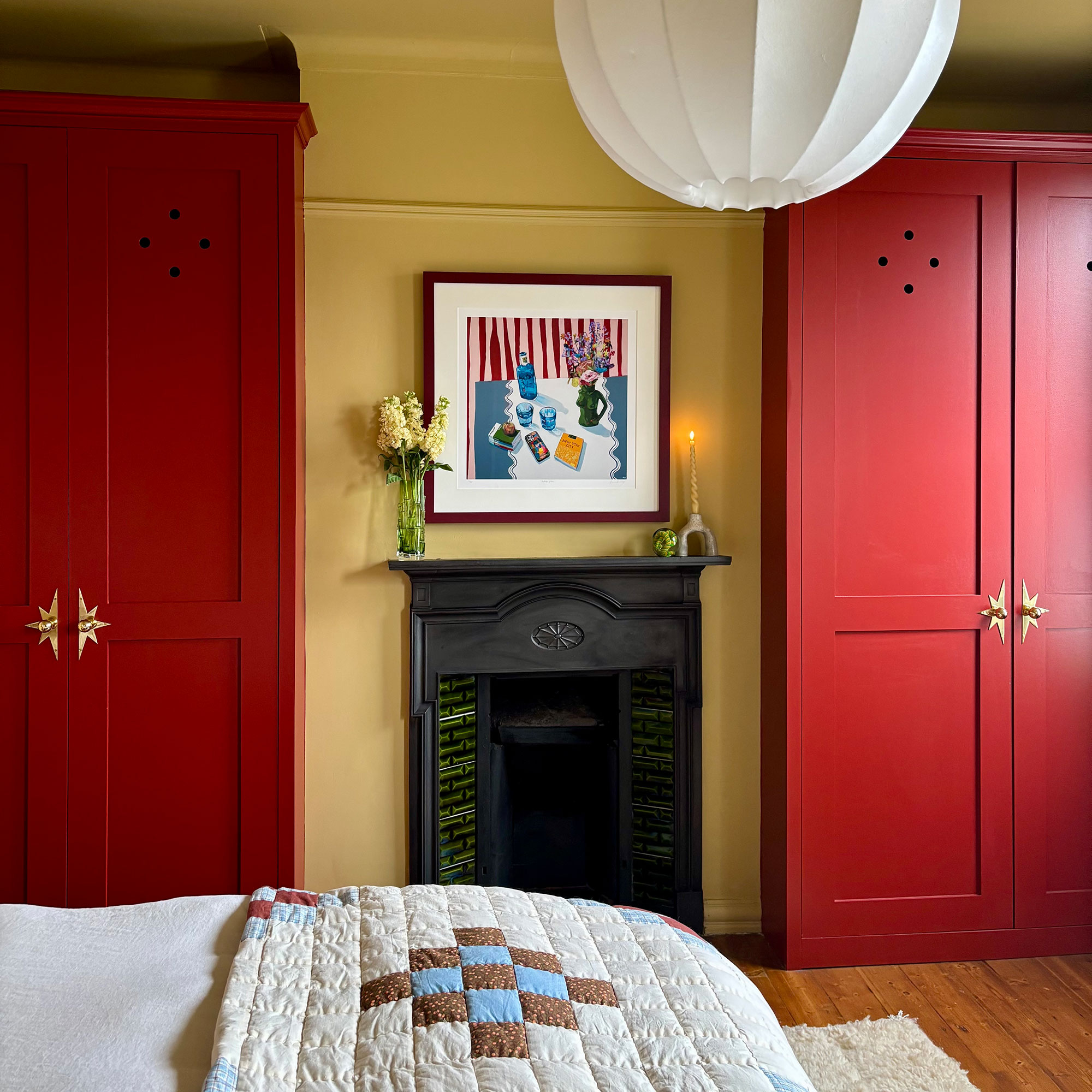 Bedroom with yellow walls and two red painted wardrobes on either side of original black iron fireplace topped with artwork