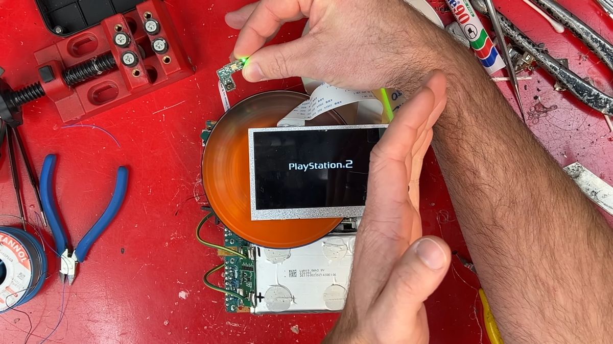 Hand holding DIY PS2 handheld power connector with disc spinning behind LCD screen and PlayStation 2 logo displayed.
