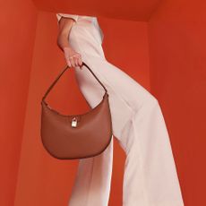 A person holding a rust-red handbag from Fiorelli in front of a red backdrop.