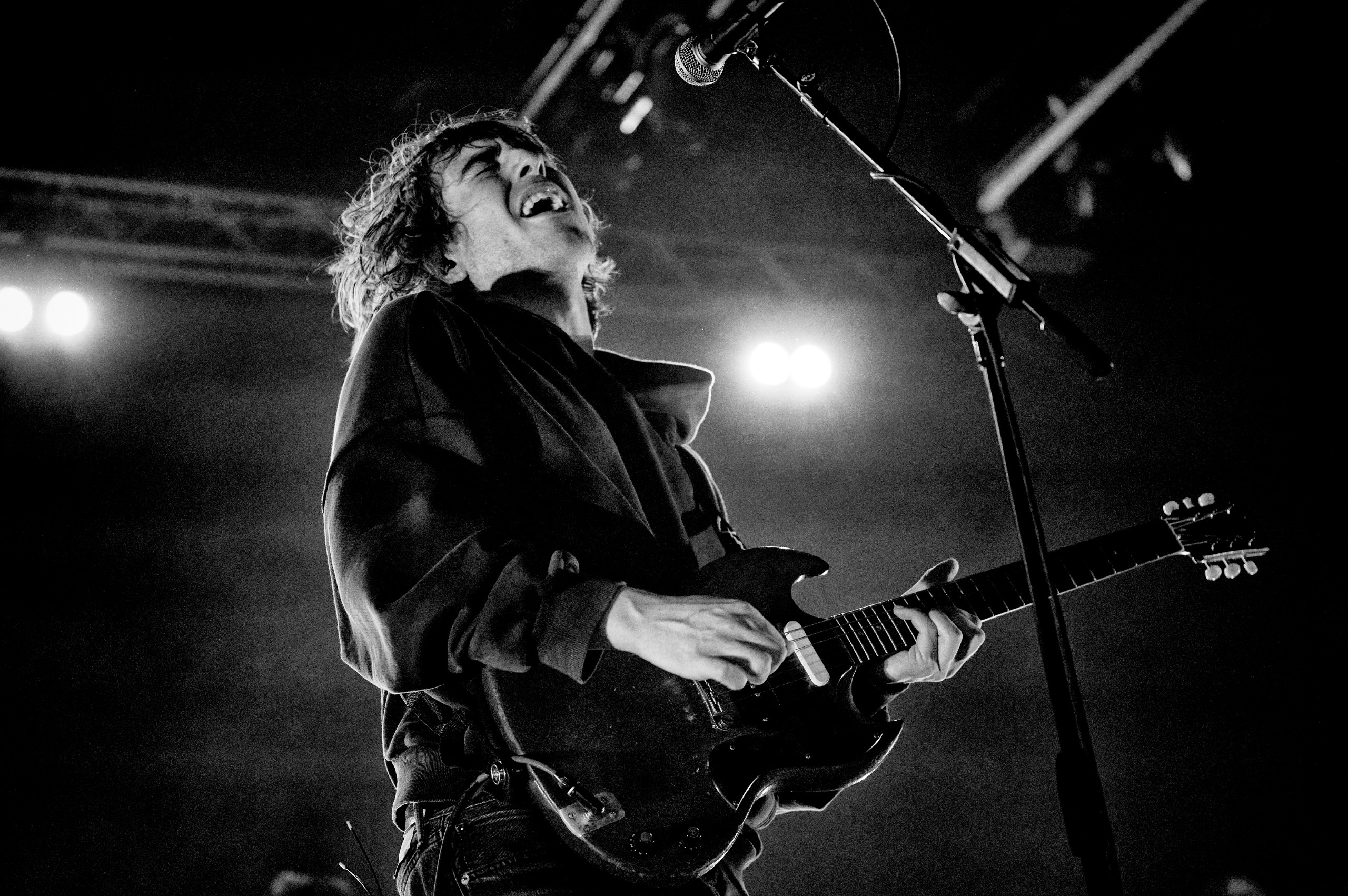 A low-angle, black-and-white photo of Cameron Winter passionately singing and playing an electric guitar onstage under bright stage lights. 