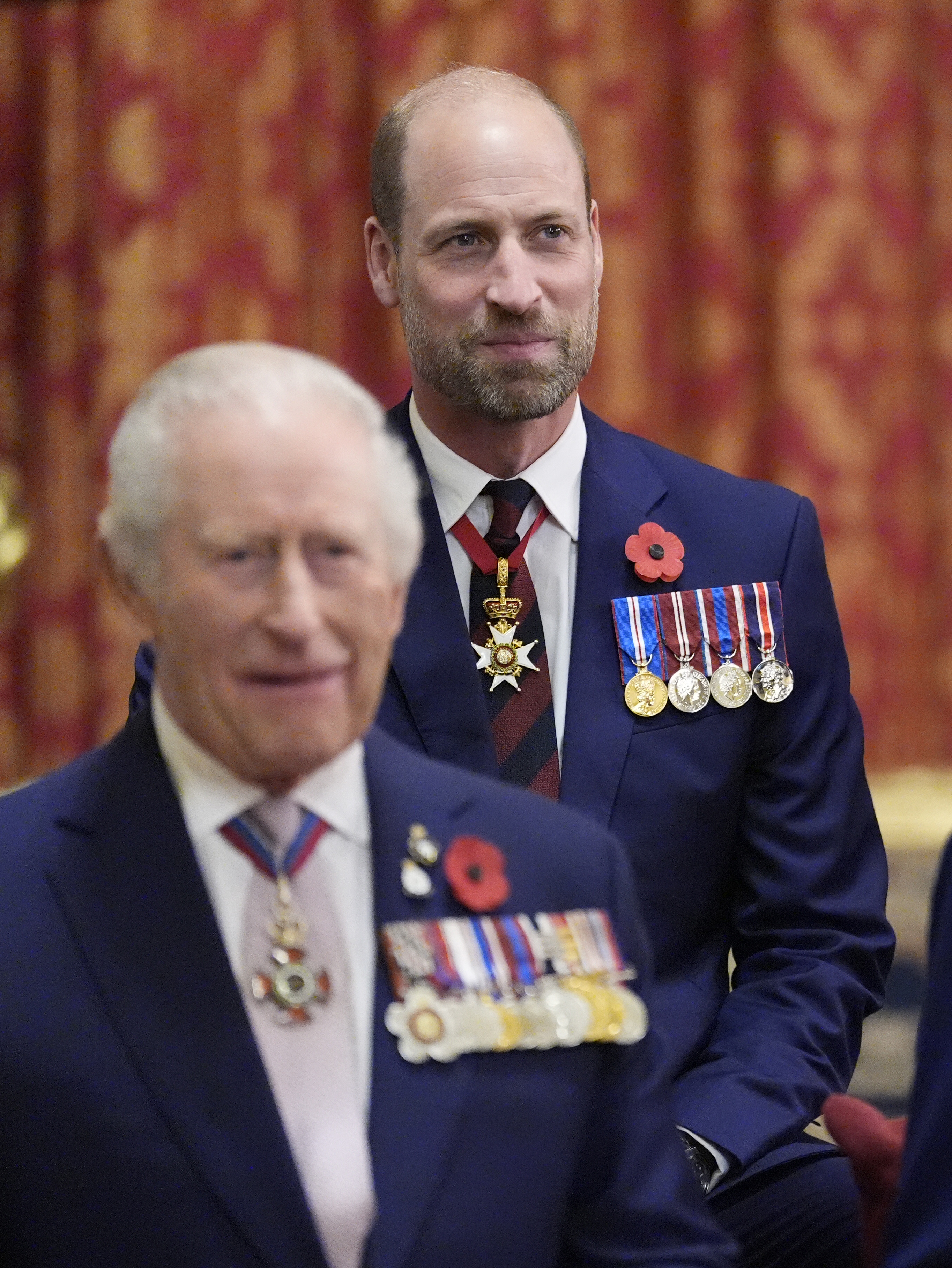 Prince William standing behind King Charles both wearing navy suits and medals