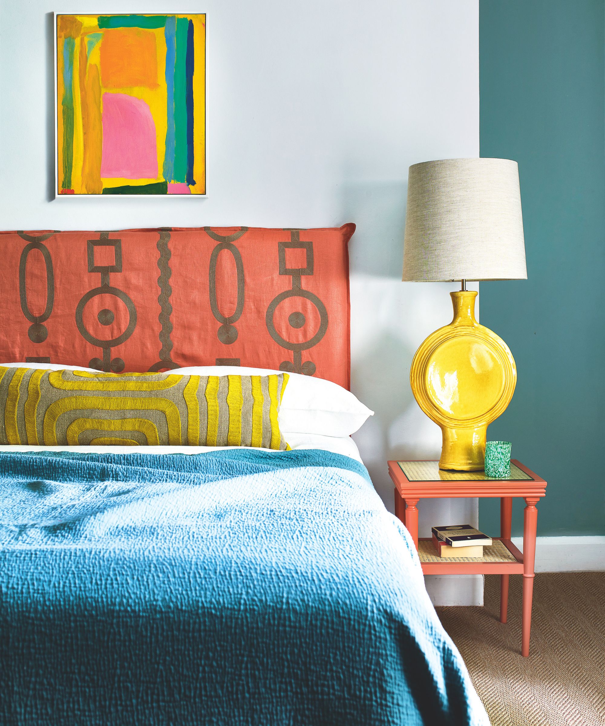 A bright and colorful bedroom with a large double bed with orange patterned headboard to the left, with white bedding, an ochre and grey patterned bolster cushion, and large blue blanket. To the right of the bed is an orange bedside table with a stack of books, yellow lamp with white lampshade, and green glass of water, and the walls behind are white and blue. Above the bed is a colorful artwork hanging in the centre, and the carpet is jute.