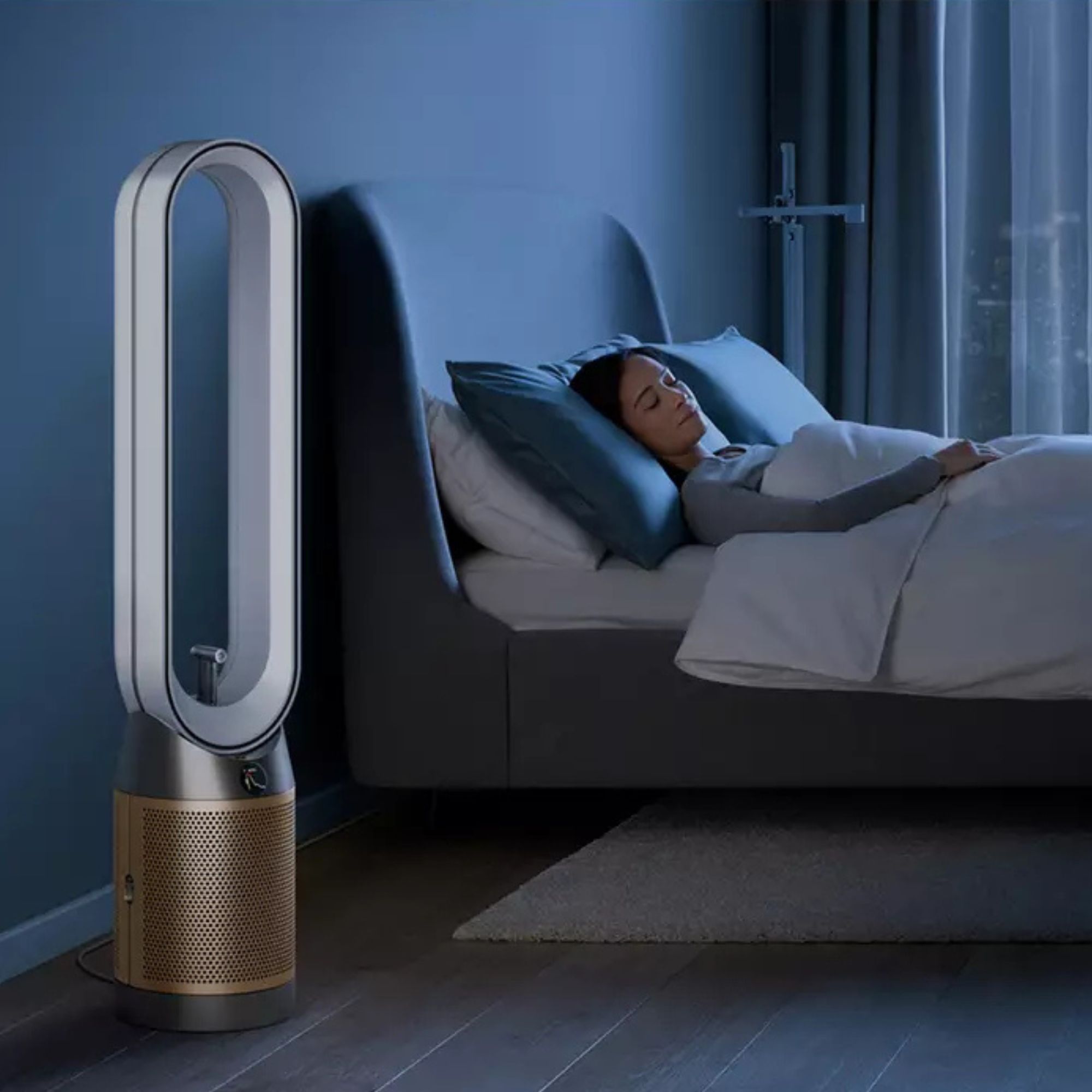 Dyson Find+Follow purifying fan sitting on the floor in a bedroom, with a woman sleeping in a bed next to it