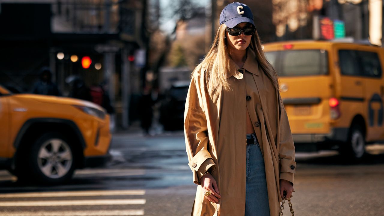 A new york attendee at fashion week fall winter 2025 wearing trench coat and navy baseball cap and sunglasses 