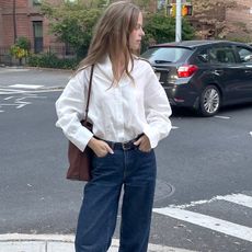 Scandinavian influencer and creative Clara Dyrhauge poses at a NYC crosswalk wearing a white linen button-down shirt, a brown leather tote bag, black belt, and relaxed dark-wash jeans