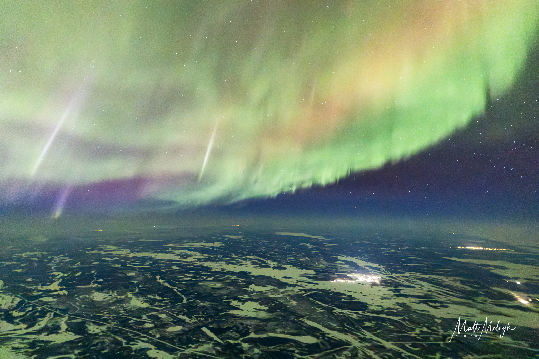 curtains of green auroras fill the sky, with broken clouds below and distant glows of city lights. The aurora is also peppered with long thin white pillars.