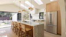Modern kitchen with large stainless steel fridge freezer, marble kitchen island with four rattan chairs, three golden hanging lights. Pictured in background is cooker, two small windows, white bricked tile splash wall, and outdoor area with table and chairs.