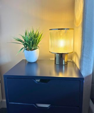 My matte navy nightstand table clear of dust and clutter, with an art deco fluted glass round gold lamp and white plant pot with greener