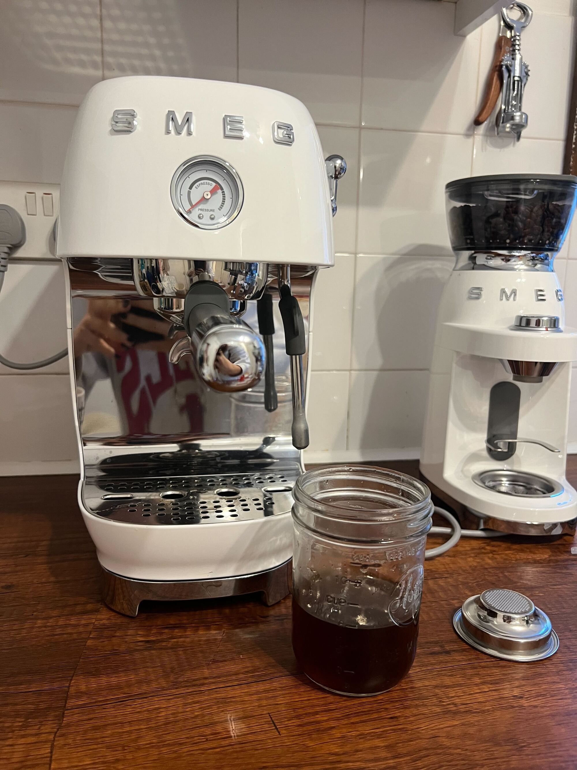 Image of a mason jar half-filled with cold brew a white Smeg cold brew machine on a wooden countertop with white tile backsplash