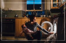 young mother holding crying baby while trying to also load washing machine in kitchen at night