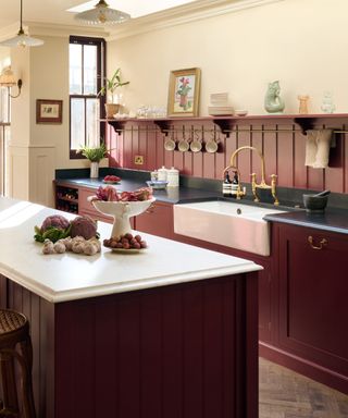 deep red kitchen with cream walls, open shelf, butler sink, matching island, brass hanging rail, fluted shade pendant lights, white countertops,