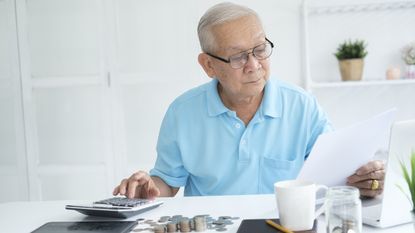 An older man looks at paperwork and uses a calculator while sitting at his desk at home.