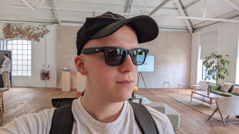 Hamish wearing a black pair of Wayfarer smart glasses from Ray-Ban and Meta. He's also wearing a hat and a bag in a large modern living room.