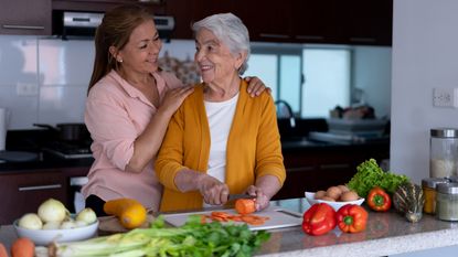 A mother cuts up vegetables as her caregiving daughter hugs her from the side.