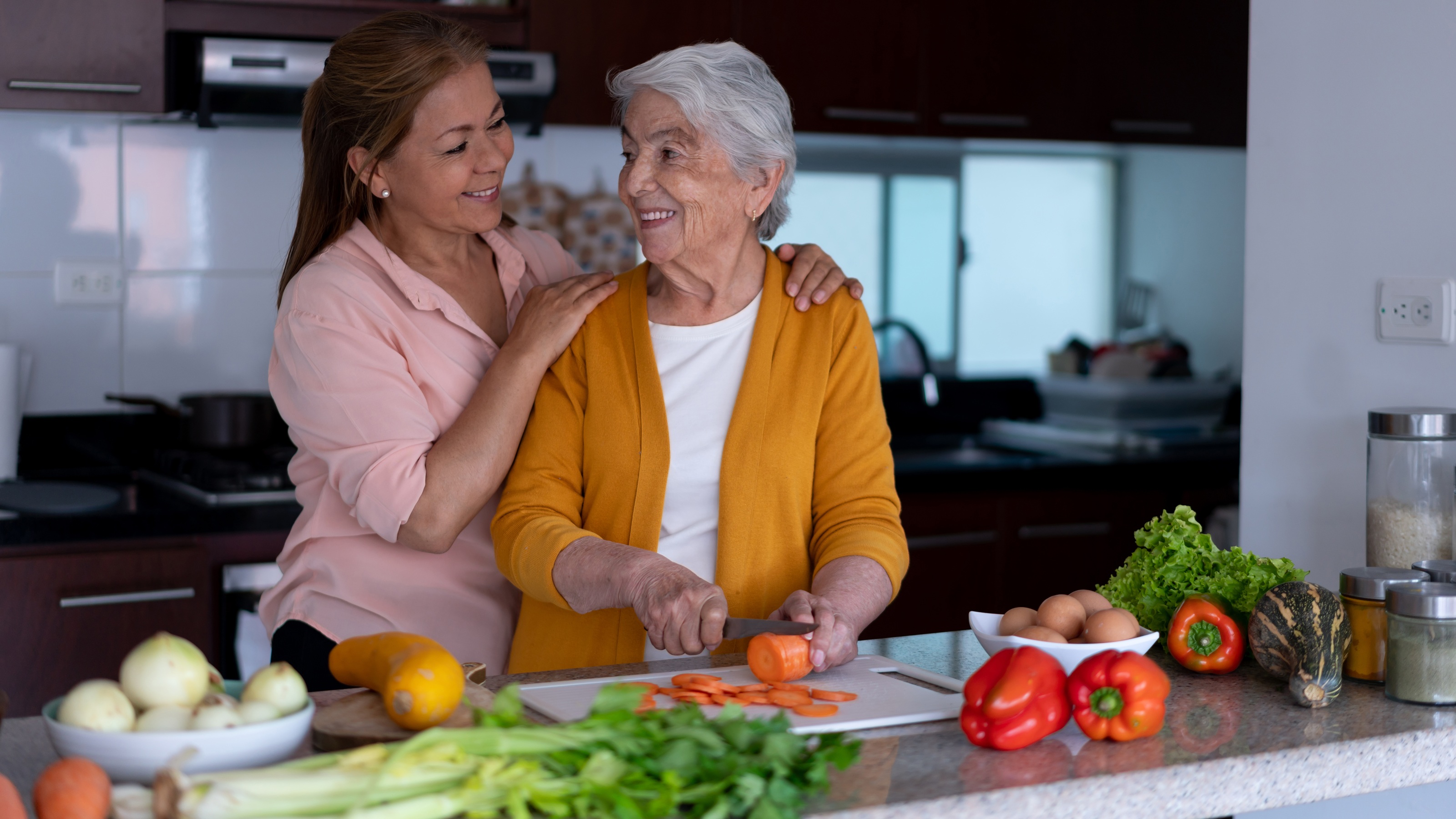 A mother cuts up vegetables as her caregiving daughter hugs her from the side.