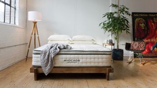 Avocado Green Mattress review: An image showing the mattress sat on a wooden bed frame, with a chunky grey knit blanket drapped across the edge. In the background sits a poster for the film Farenheit 45