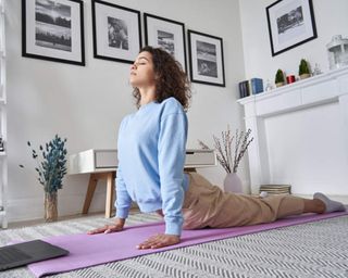 A female wearing a pastel blue sweatshirt taking part in a virtual yoga session with pink-purple yoga mat in living room