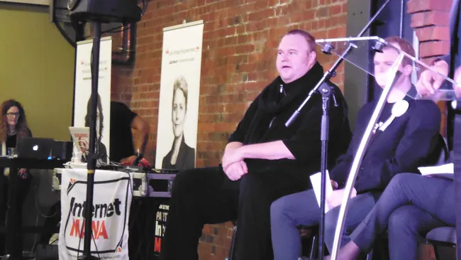 La imagen muestra a Kim Dotcom sentado en un panel de discusión, posiblemente en un evento o conferencia. Está vestido de negro y parece estar hablando o escuchando atentamente. A su alrededor hay otras personas sentadas, y en el fondo se observan una pared de ladrillo y algunos carteles, uno de los cuales parece tener la imagen de una persona. Delante de Dotcom, hay una mesa con un cartel que dice "Internet Mana", posiblemente el nombre de la organización o el evento.