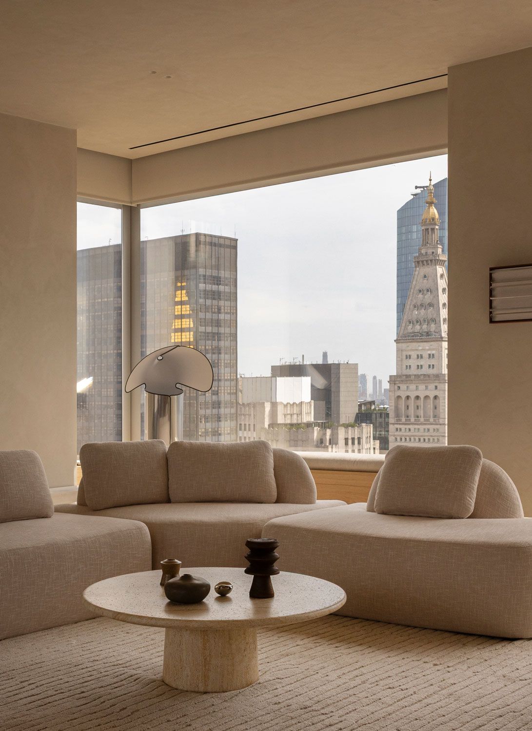Neutral-toned living room at 262 Fifth Avenue with modular seating, a round stone coffee table and expansive windows framing Manhattan architecture