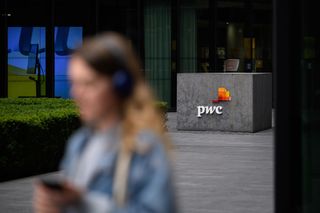 The a sign with the letters PwC displayed outside of an office building, with a blurred woman walking in the foreground