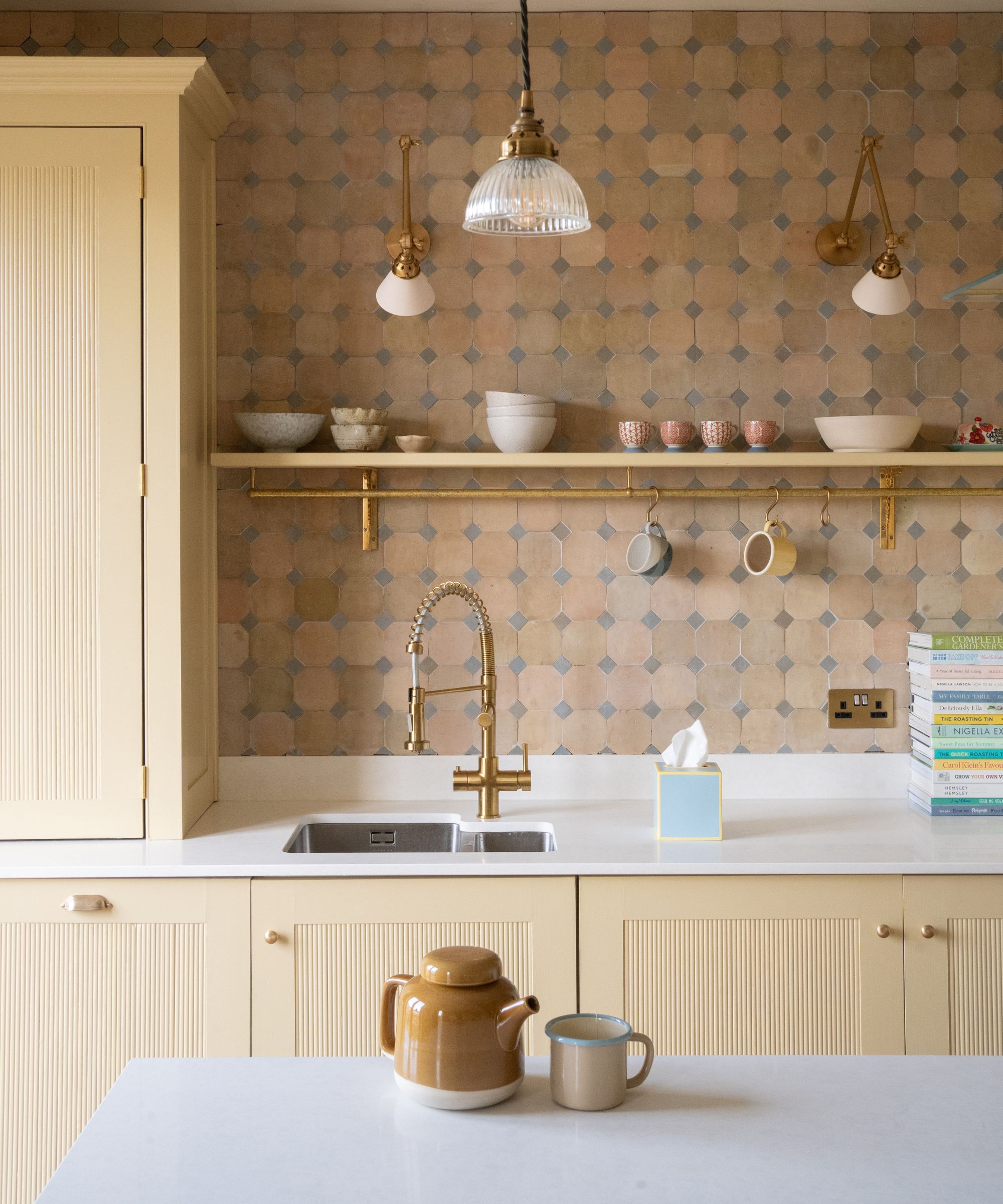 Soft cream kitchen with fluted cabinet fronts and a white worktop. A brass tap and matching wall lights sit against a geometric tiled splashback in muted beige and blue