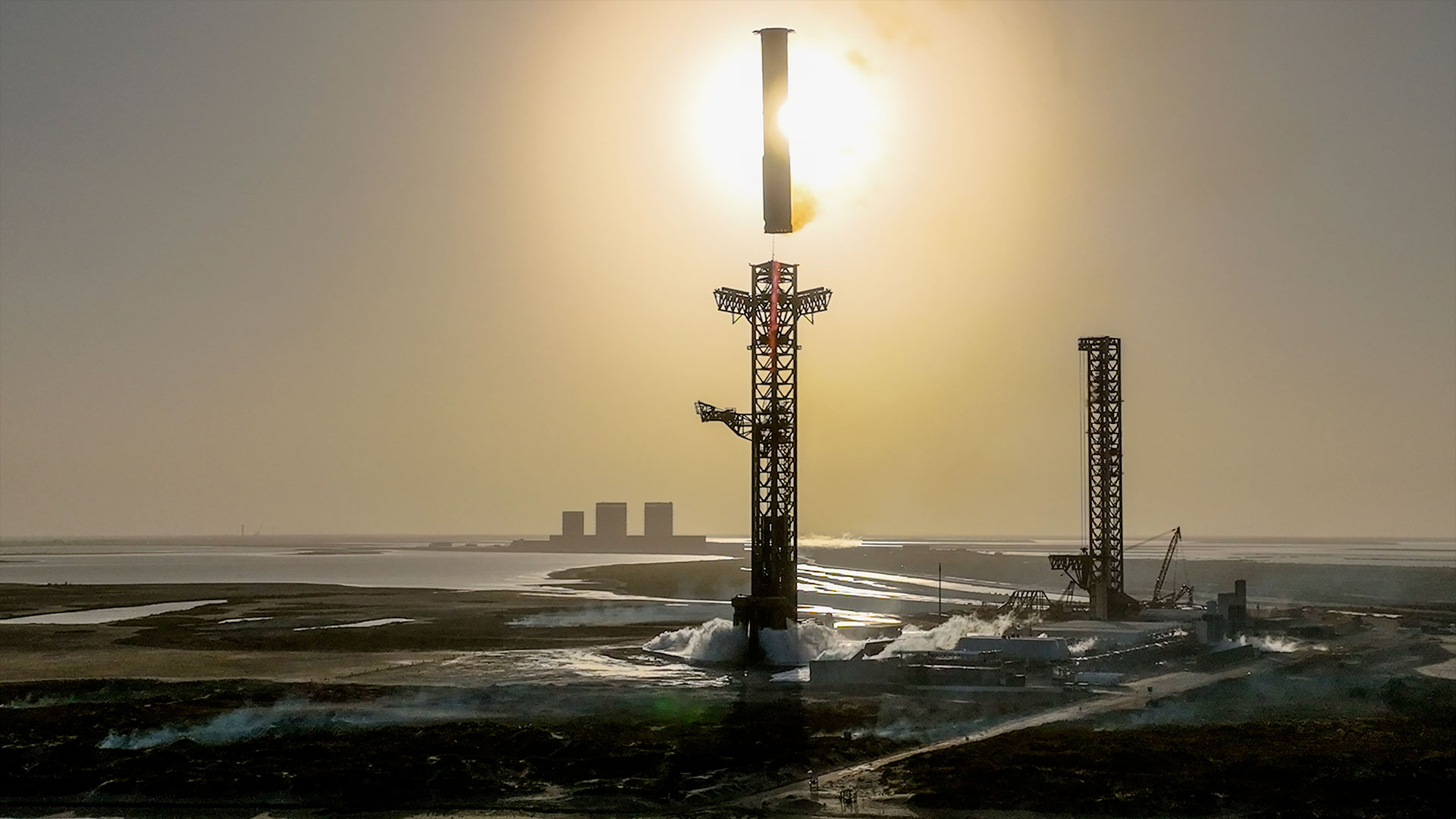 a rocket booster approaches its launch tower to be caught as both are silhouetted by the sun