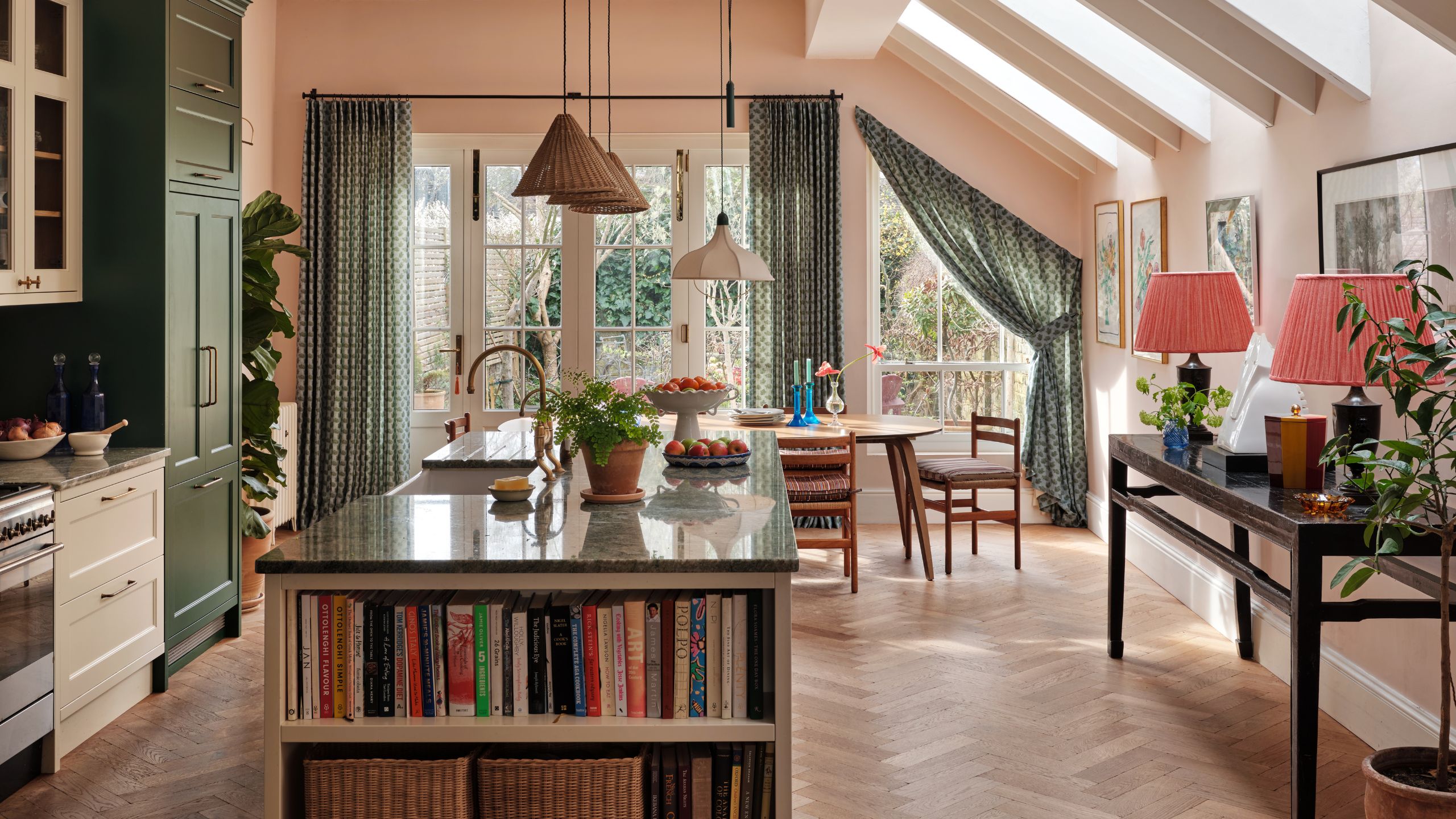 a large extended London kitchen with soft pink walls, deep gren and white two tone cabinetry, herringbone floors, pendants and dining area framed by drapes at the far end