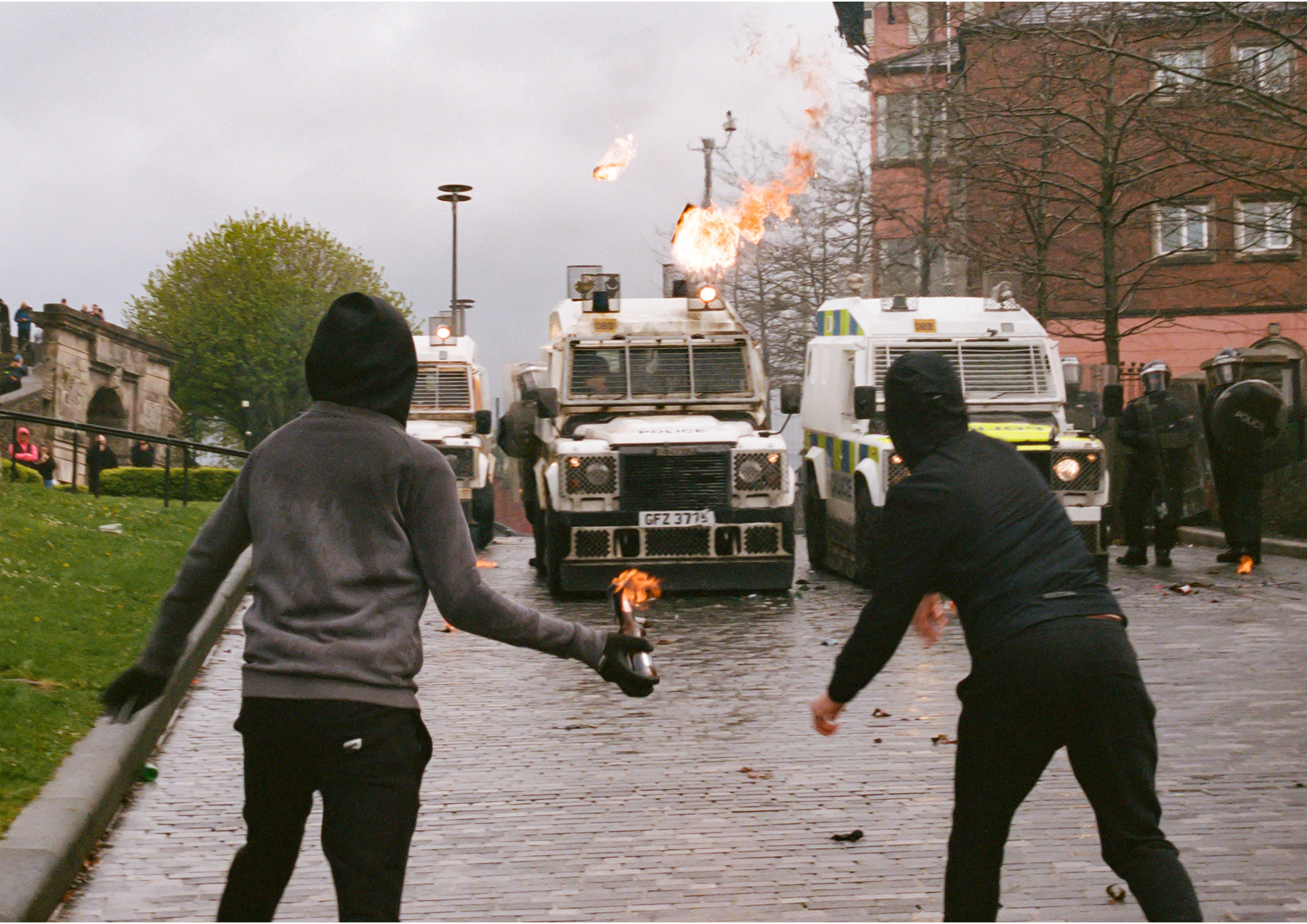 Two individuals in dark hoodies and masks throw lit Molotov cocktails toward a line of armored police Land Rovers on a cobblestone street.