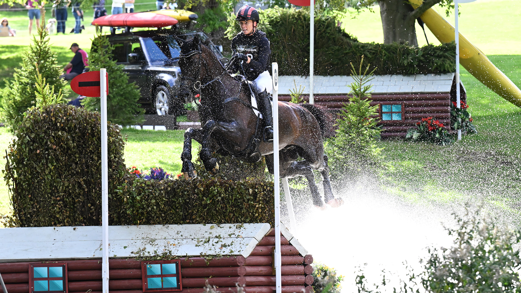 Caroline Pamukcu, a rider in a black top with a hat with black and red stripes, pilots HSH Blake, a dark horse, over a brush fence in water at Burghley