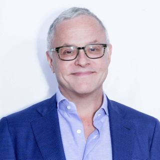 A headshot of Neal Baer, a middle aged white man wearing black rimmed glasses and a blue suit and button down shirt