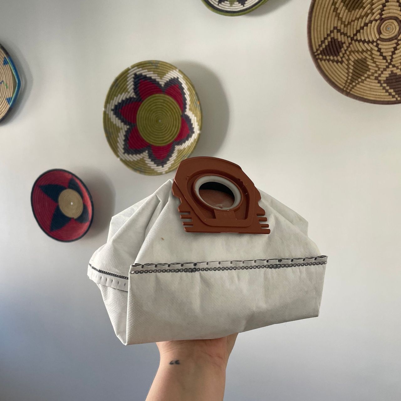 Holding a Miele vacuum cleaner bag in front of a white wall with colourful baskets on it