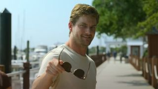 Jake Weary in a tight t-shirt standing on a doc and smirking while holding sunglasses in The Waterfront.