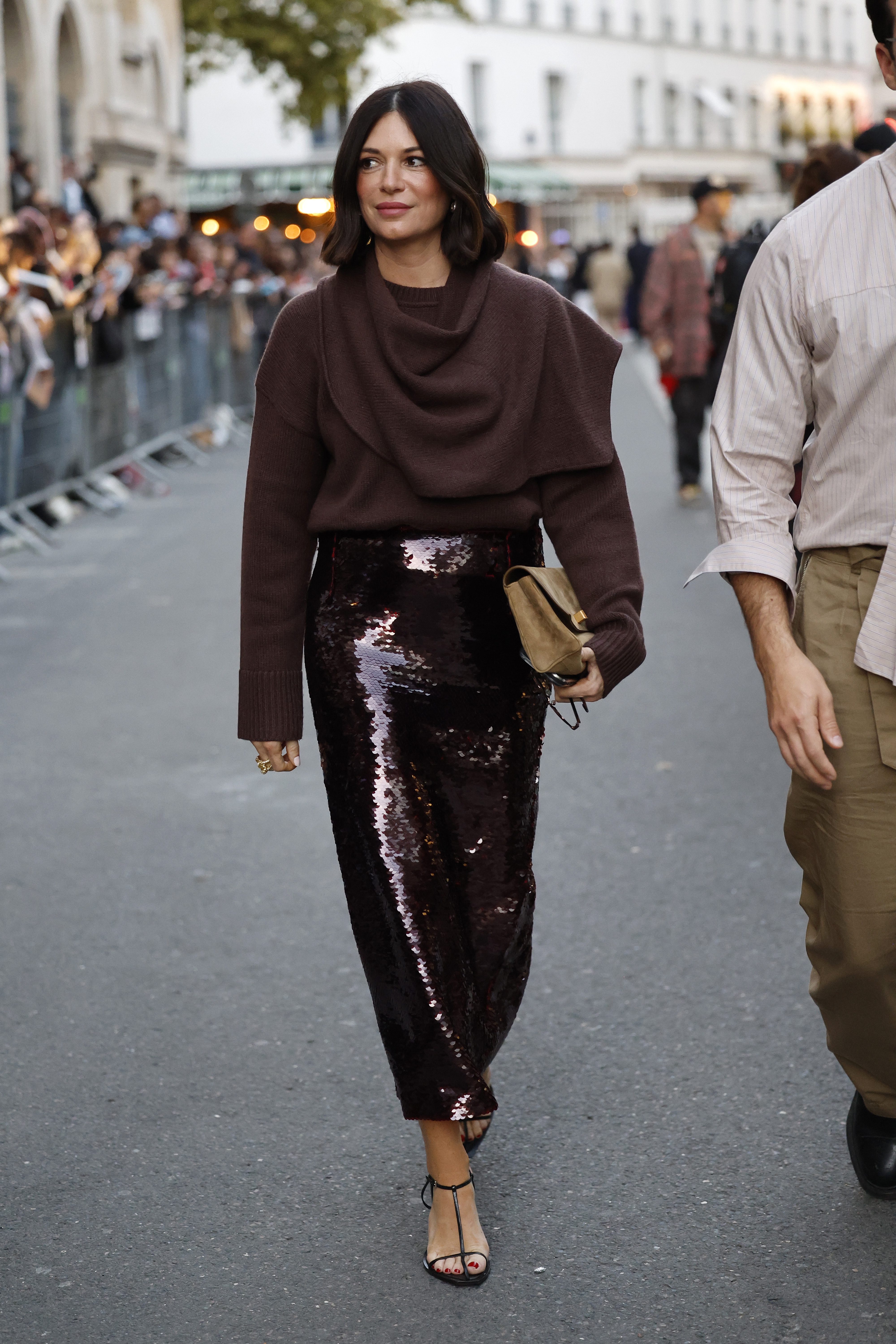 A woman wears a brown sweater, burgundy sequin skirt, suede clutch, and sandals.