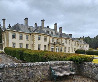 A white brick mansion with a stone wall to the side of the house and a bench and a hedge