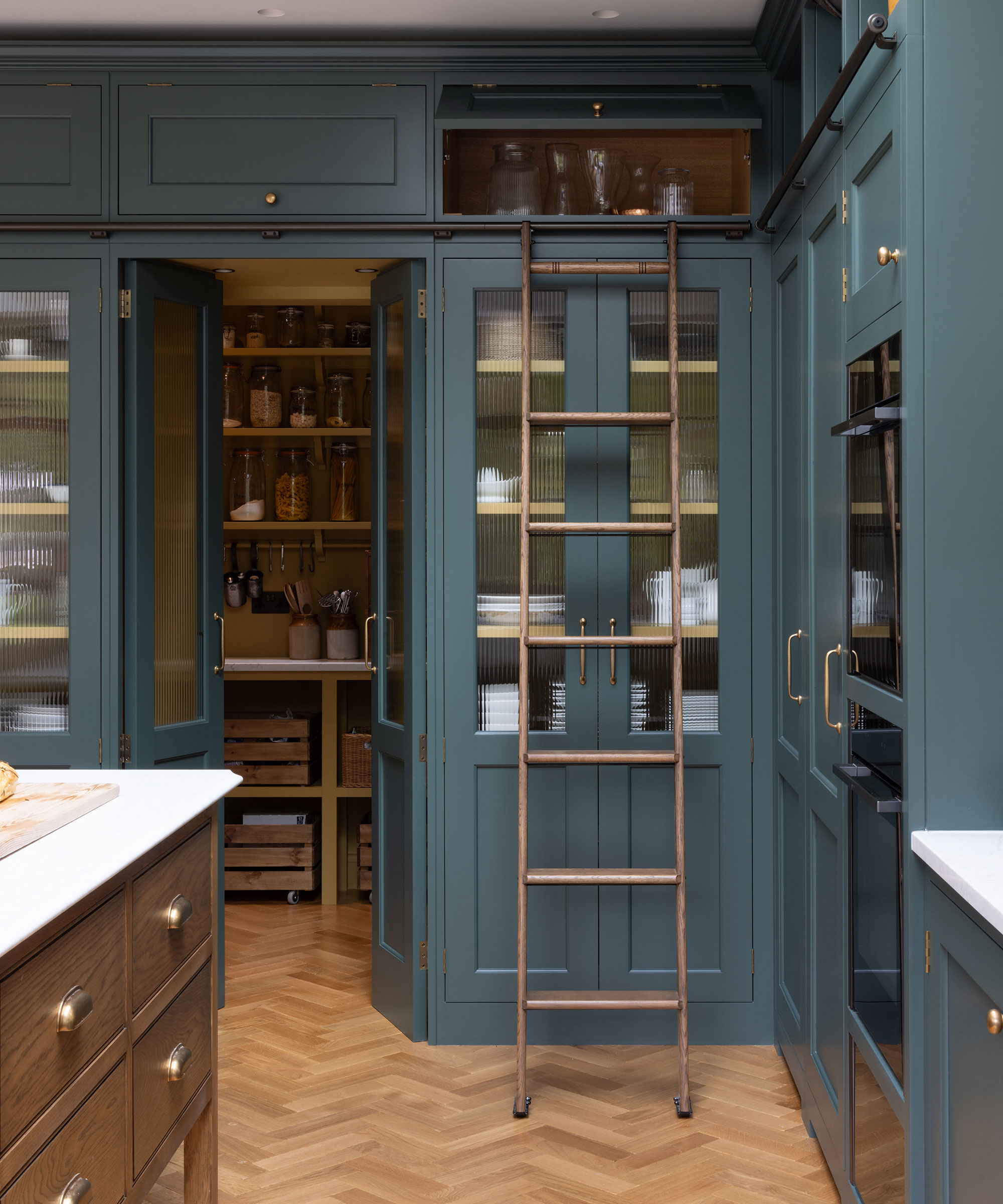 blue Shaker kitchen with brass handles and herringbone flooring with ladder to reach tall units