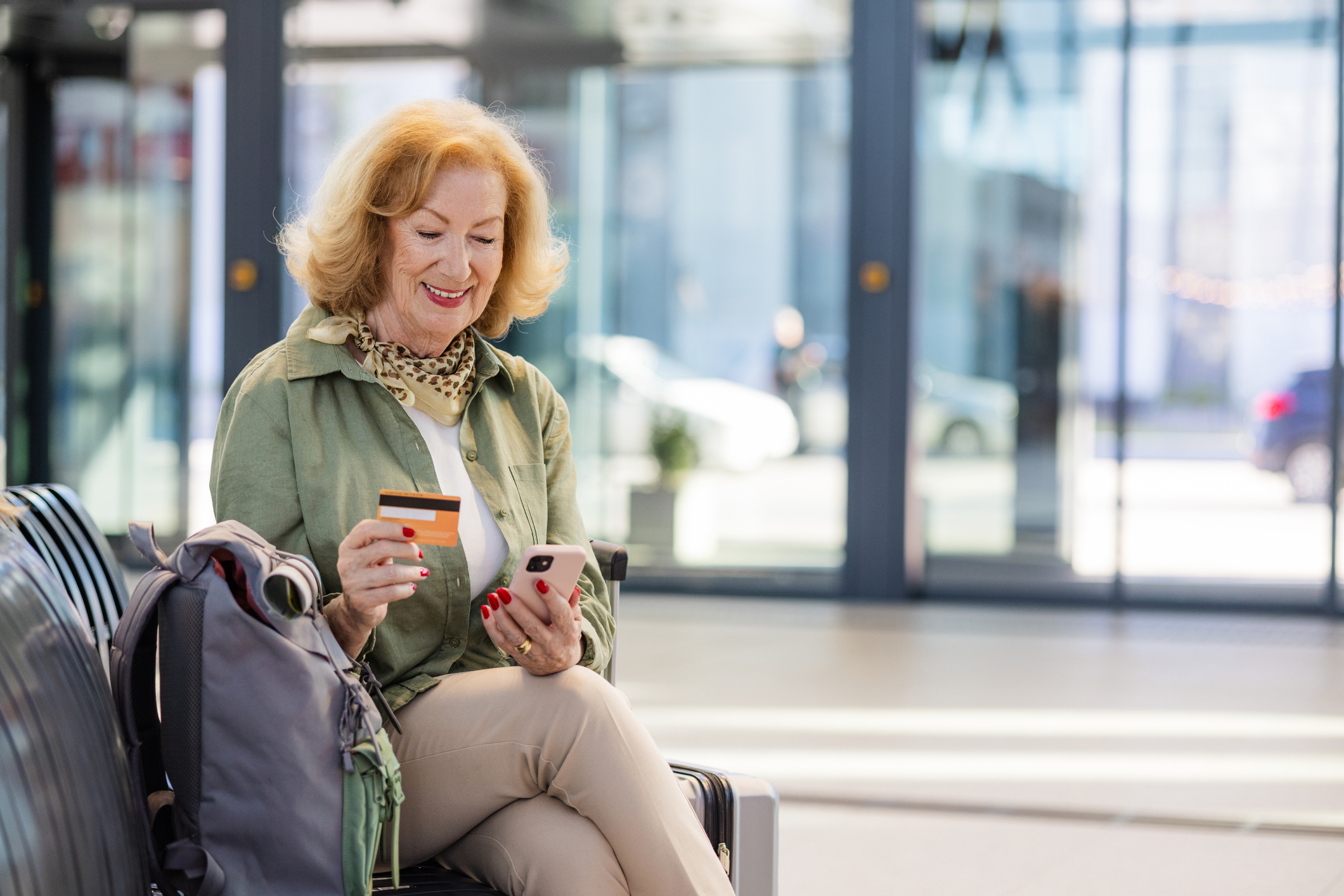 A senior woman sits at an airport while using her credit card and phone.