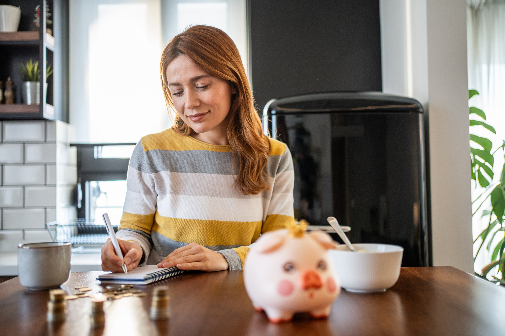 woman sitting at kitchen table writing down expenses in notebook, managing home budget, adding to savings and investments