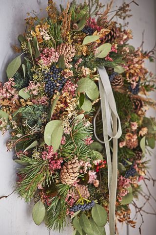 A Christmas wreath made of dried and fresh foliage, seed heads and pinecones, green leaves, black berries and silver ribbons.