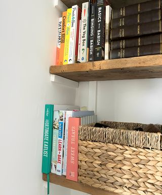 Bright cookbooks on wooden plank shelves next to leather bound books and a woven storage basket