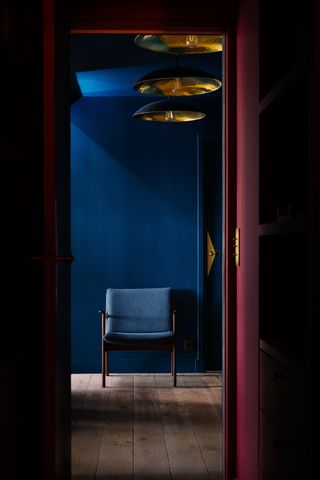 Image of a hallway that is painted in a deep red color with a deep blue wall at the end of the hall. There is a blue accent chair at the end of the hall as well. Three gold pendant lights line the hallway.